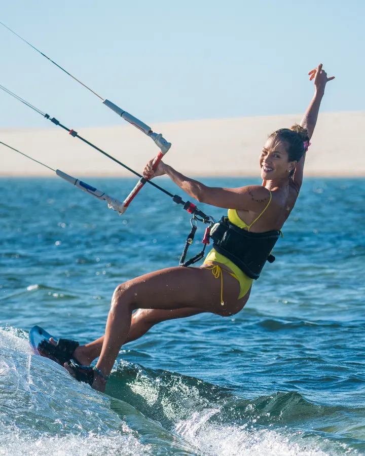 Eugênia velejando de kite, com um braço para o alto e as dunas ao fundo
