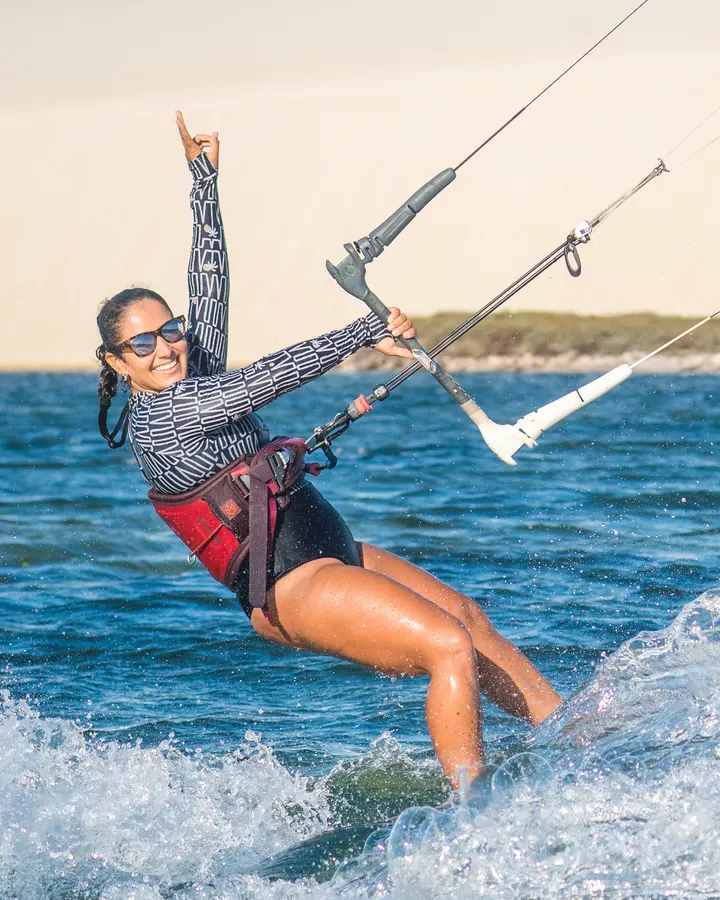 Itana velejando de kite, com um braço para o alto e as dunas ao fundo