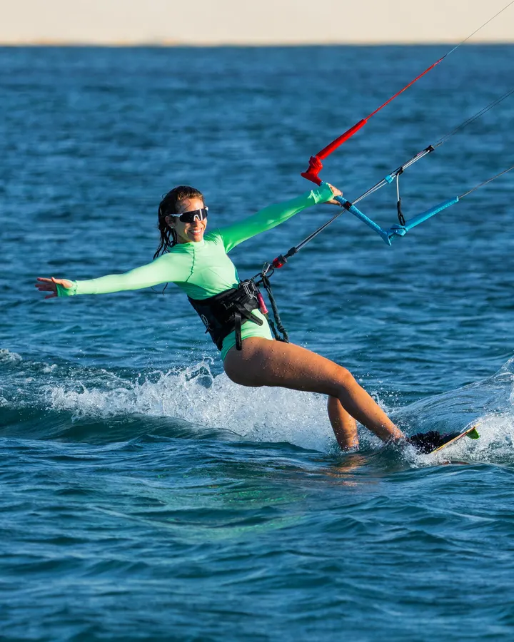 Maria Amélia velejando de kite, de braços abertos, com as dunas ao fundo