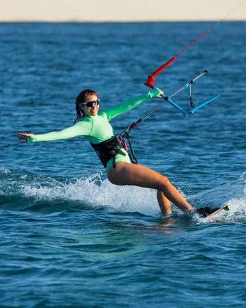 Maria Amélia velejando de kite, de braços abertos, com as dunas ao fundo